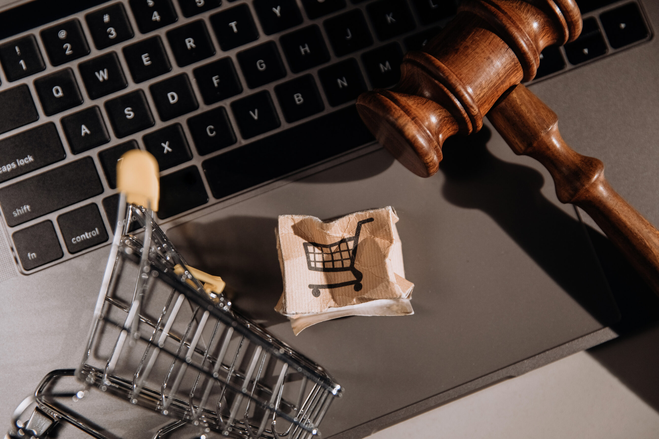 Symboliczne ujęcie sprzedaży online — zgniecione pudełko z ikoną koszyka, miniaturowy wózek i młotek sędziowski na tle klawiatury, podkreślające znaczenie praw konsumenta w e-commerce.